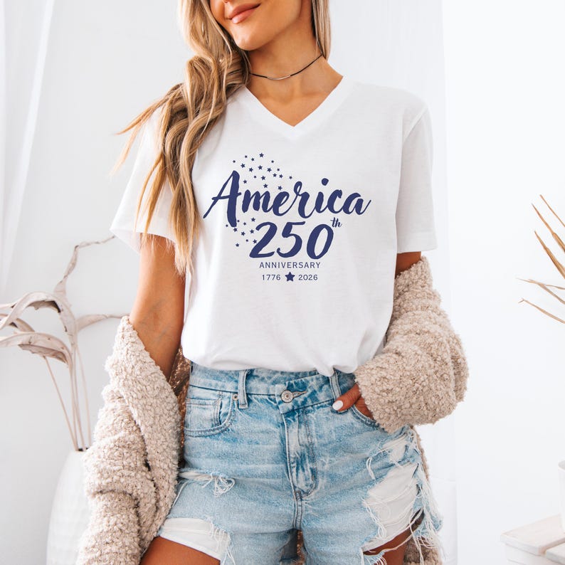 May include: A white short-sleeved V-neck t-shirt featuring dark blue text that reads America 250th Anniversary 1776-2026, accompanied by a cluster of small stars and a single star icon between the years, printed on the front of the garment.
