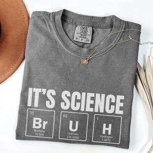 May include: A gray t-shirt with the words "IT'S SCIENCE" printed in white. Below the text are three periodic table elements: Bromine, Uranium, and Hydrogen. A silver necklace with a pendant is draped across the shirt.