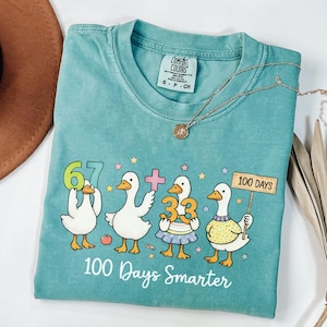 May include: Teal Comfort Colors t-shirt with a graphic of four cartoon ducks holding numbers and a sign that reads "100 Days." The text "100 Days Smarter" is below the ducks. A gold necklace is draped on the shirt.