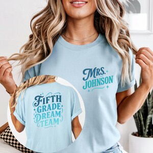 May include: Light blue t-shirt with the text "Mrs. Johnson" in dark blue script and light blue accents. The back of the shirt features the text "Fifth Grade Dream Team" in a similar color scheme. The shirt is a crew neck style.