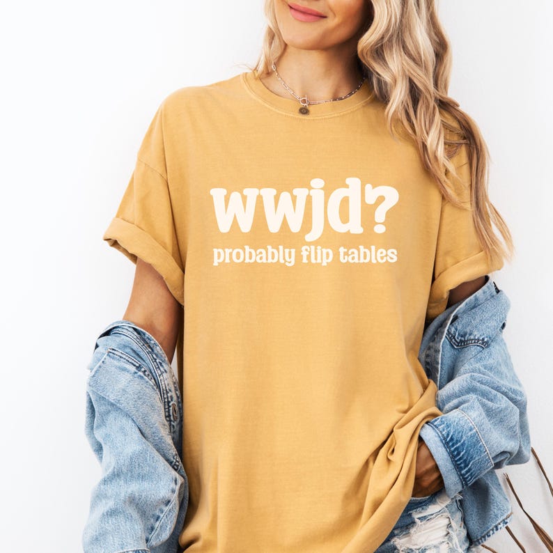 May include: Mustard yellow t-shirt with the text "wwjd? probably flip tables" in white. The shirt has rolled-up sleeves and is paired with a denim jacket. The shirt is a casual wear item.