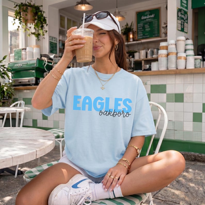 K&ouml;nnte beinhalten: Ein hellblaues Kurzarm-T-Shirt mit dem Wort EAGLES in gro&szlig;en blauen Blockbuchstaben und dem Wort oakboro in schwarzer Schreibschrift darunter. Das T-Shirt wird mit einer wei&szlig;en Baseballkappe und wei&szlig;en Sportschuhen getragen.
