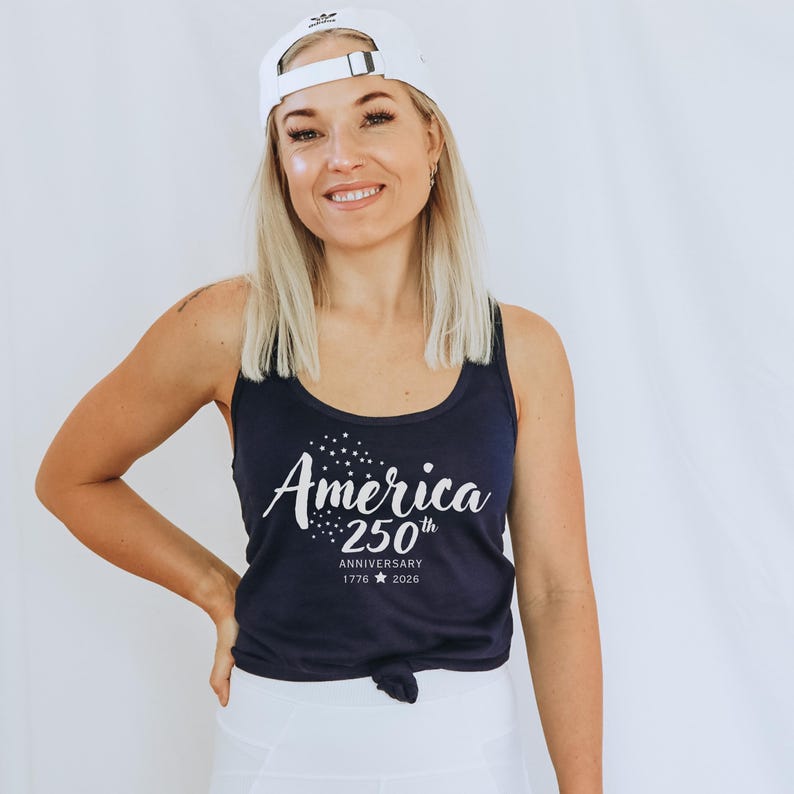 Pu&ograve; includere: Una canotta blu navy con la scritta "America 250th Anniversary 1776 2026" in bianco. La persona indossa un berretto da baseball bianco.