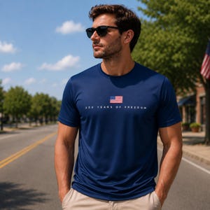 May include: A navy blue short-sleeved t-shirt featuring a small American flag graphic centered on the chest above the white text 250 YEARS OF FREEDOM. The shirt has a crew neckline and is worn with light-colored trousers.