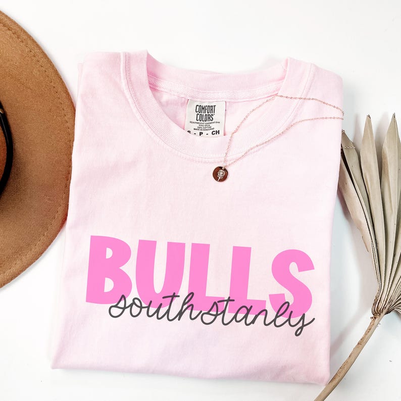 K&ouml;nnte beinhalten: Ein hellrosa T-Shirt mit kurzen &Auml;rmeln, das das Wort BULLS in gro&szlig;en, fetten rosa Buchstaben zeigt, mit dem Text south stanly darunter in einer dunkelgrauen Schreibschrift. Das T-Shirt hat ein Comfort Colors Markenetikett am Innenkragen.