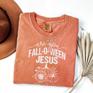 May include: An orange Comfort Colors t-shirt with the text "are you FALL-O-WEEN JESUS" in white. The shirt features a graphic of pumpkins and sunflowers. A gold necklace and a brown hat are also visible.