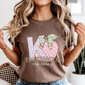 May include: A brown t-shirt featuring a colorful graphic design. The design includes the letters "K" and "6" with floral and checkered patterns, a bow, and a pencil. The words "Kindergarten mrs. honey" are written below the graphic.
