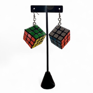 Puede incluir: Un par de pendientes de cubo de Rubik. Un cubo presenta la combinación de colores clásica de rojo, naranja, amarillo, verde, azul y blanco. El otro cubo tiene una paleta de colores apagados. Ambos cubos están suspendidos de ganchos plateados.