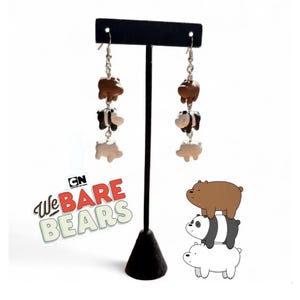May include: Dangle earrings featuring three cartoon bear figures in brown, black and white. The earrings are displayed on a black stand. The image also includes the text "We Bare Bears" and the CN logo.