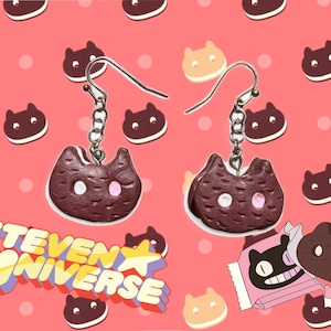 May include: A pair of dangle earrings featuring a cat-shaped cookie design. The earrings are dark brown with white and pink accents, hanging from silver-colored hooks. The background is pink with repeating cat cookie graphics and the text "STEVEN UNIVERSE".