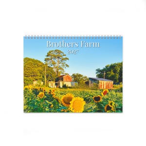 May include: A spiral-bound wall calendar titled Brothers Farm 2027. The cover features a landscape photograph of a field of bright yellow sunflowers in the foreground with rustic farm buildings and green trees under a clear blue sky.