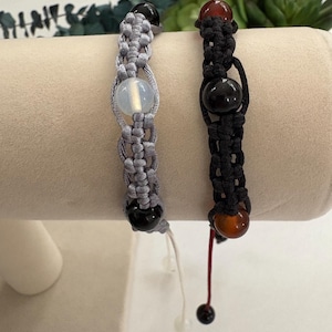 May include: Two macrame bracelets with gemstone beads. One bracelet is gray with a white and black bead, the other is black with brown and black beads. The bracelets are displayed on a beige stand.