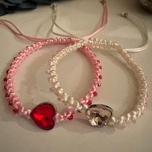 May include: Two macrame bracelets. One is pink and white with a red heart-shaped gem. The other is white and pink with a clear, faceted gem. Both bracelets have small red beads and adjustable closures.