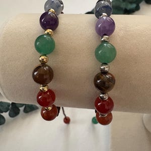 May include: Two beaded bracelets with a variety of colorful gemstones. The bracelets feature a sequence of round beads in shades of blue, purple, green, brown, and red. Gold and silver-colored spacers separate the beads. The bracelets are displayed on a neutral background.