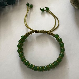 May include: A green beaded bracelet with a macrame design. The bracelet features small, round, green beads strung together with a matching green cord. The bracelet has adjustable cords with green beads at the ends.