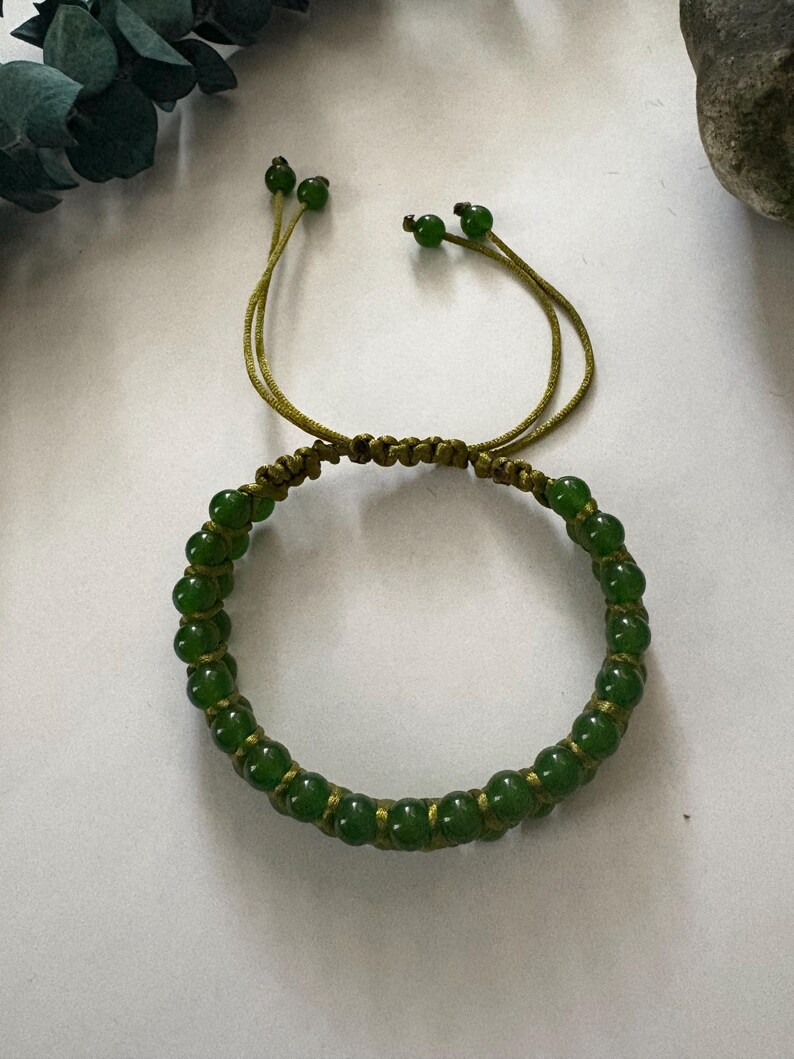 May include: A green beaded bracelet with a macrame design. The bracelet features small, round, green beads strung together with a matching green cord. The bracelet has adjustable cords with green beads at the ends.