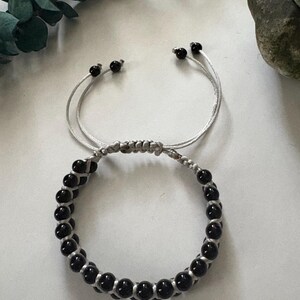May include: A black beaded bracelet with a silver cord. The bracelet features a series of small, round black beads strung together with a silver cord. The bracelet has adjustable sliding knots for a custom fit.