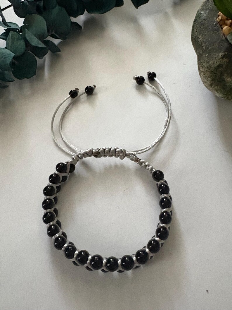 May include: A black beaded bracelet with a silver cord. The bracelet features a series of small, round black beads strung together with a silver cord. The bracelet has adjustable sliding knots for a custom fit.