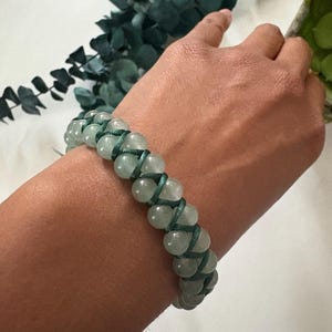 May include: A beaded bracelet featuring light green, round beads woven together with dark green cord. The bracelet is shown on a person's wrist, with a white background and green foliage in the background.