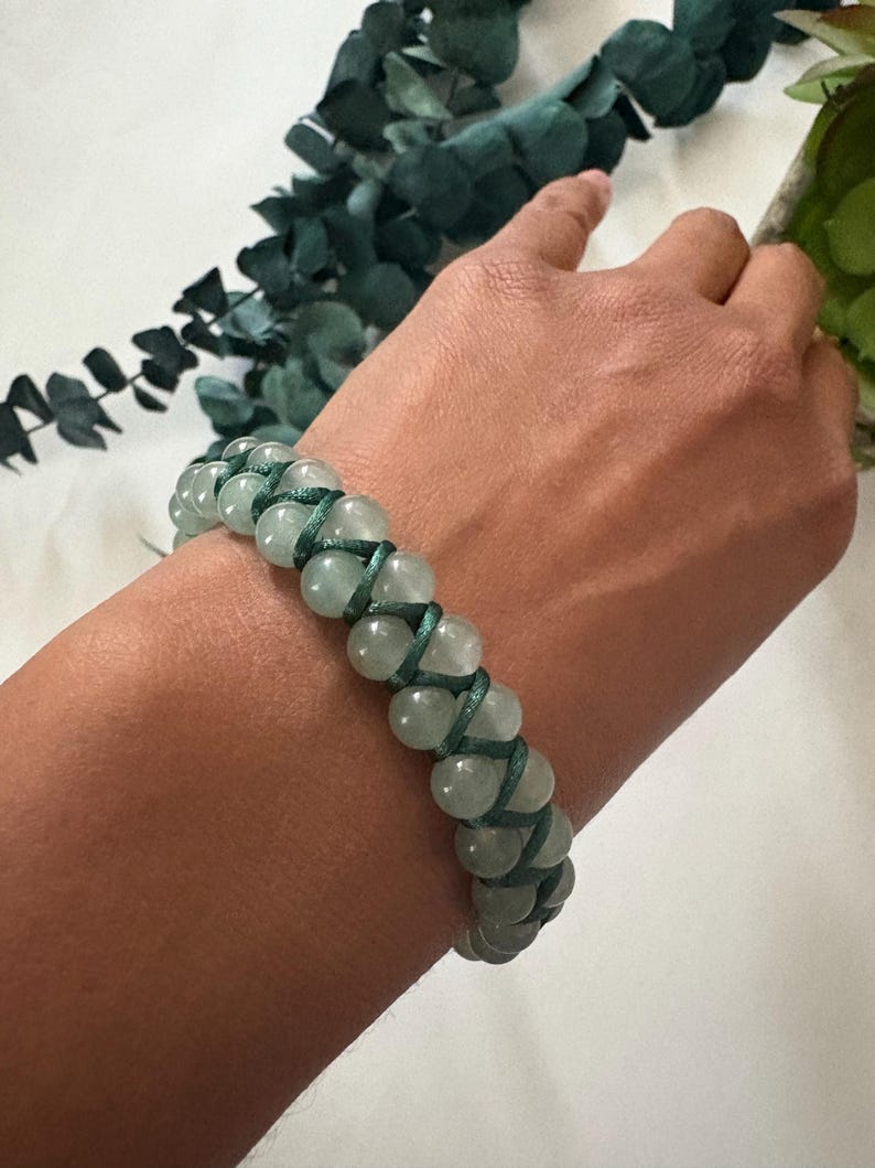 May include: A beaded bracelet featuring light green, round beads woven together with dark green cord. The bracelet is shown on a person's wrist, with a white background and green foliage in the background.