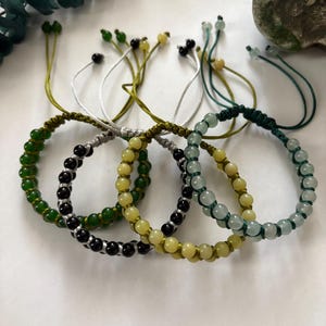 May include: A collection of beaded bracelets in various colors, including green, black, and yellow. Each bracelet features a series of small, round beads strung on a cord, with adjustable ties. The bracelets are displayed on a white surface.