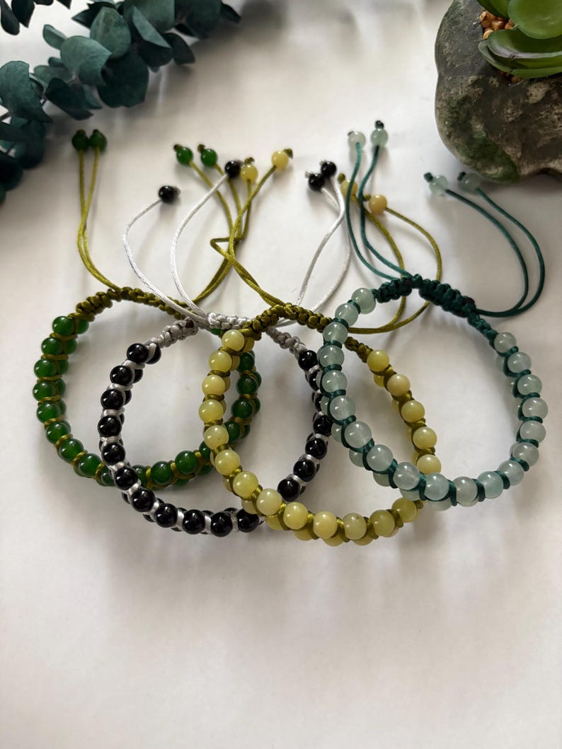 May include: A collection of beaded bracelets in various colors, including green, black, and yellow. Each bracelet features a series of small, round beads strung on a cord, with adjustable ties. The bracelets are displayed on a white surface.