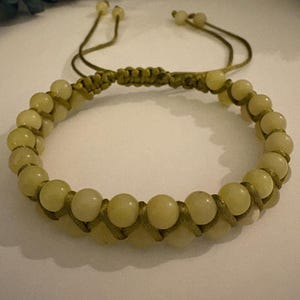 May include: A beaded bracelet featuring pale yellow-green beads. The bracelet is strung on a matching green cord, with a macrame knot design. The adjustable closure uses two cords with small beads at the ends. The bracelet is a piece of jewelry.