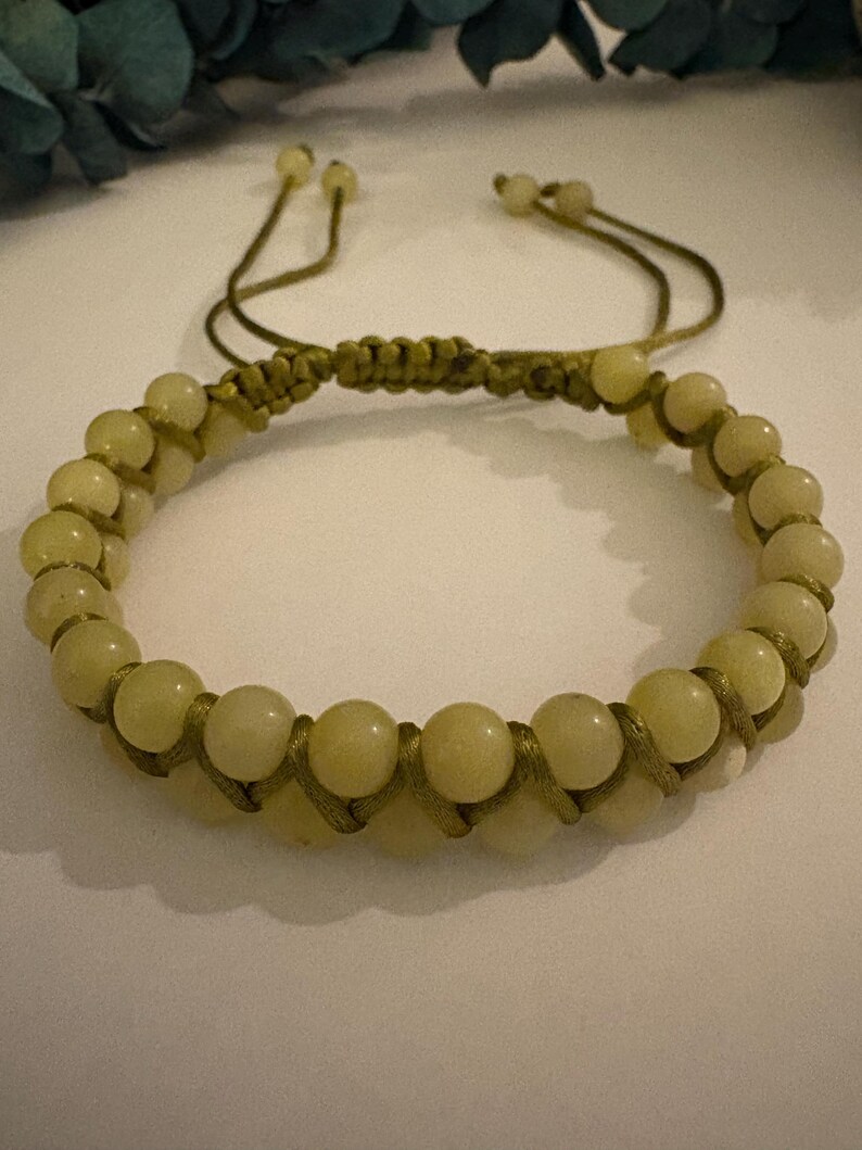 May include: A beaded bracelet featuring pale yellow-green beads. The bracelet is strung on a matching green cord, with a macrame knot design. The adjustable closure uses two cords with small beads at the ends. The bracelet is a piece of jewelry.