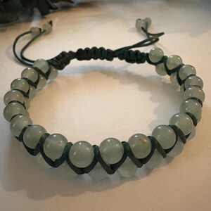 May include: A beaded bracelet featuring pale green, round beads. The beads are strung on a dark green cord, with a macrame design. The bracelet has adjustable sliding knots for a custom fit. The bracelet is a piece of jewelry.