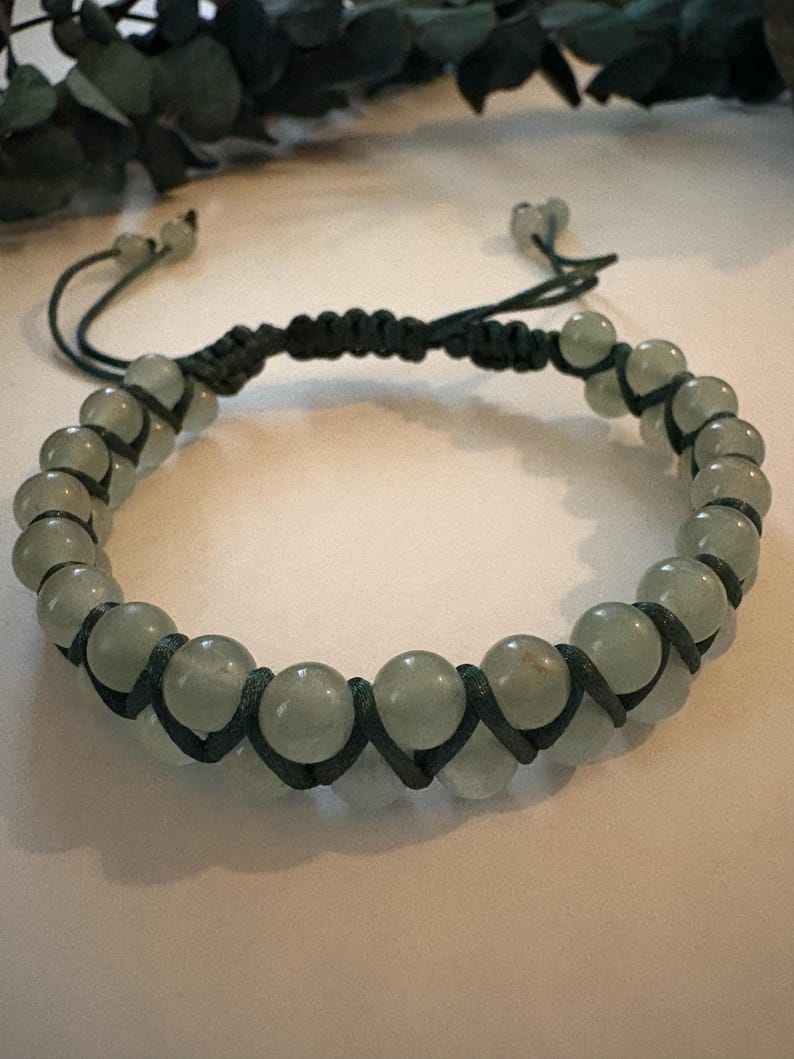 May include: A beaded bracelet featuring pale green, round beads. The beads are strung on a dark green cord, with a macrame design. The bracelet has adjustable sliding knots for a custom fit. The bracelet is a piece of jewelry.