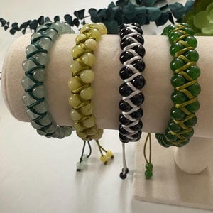 May include: Four beaded bracelets in shades of green, yellow, black, and white are displayed on a cream-colored stand. Each bracelet features a woven design with adjustable ties. The beads are round and smooth.