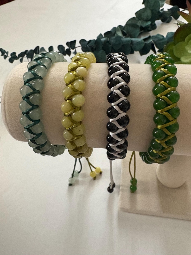 May include: Four beaded bracelets in shades of green, yellow, black, and white are displayed on a cream-colored stand. Each bracelet features a woven design with adjustable ties. The beads are round and smooth.