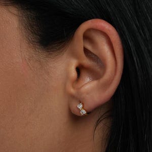 May include: A close-up of an ear with a small gold hoop earring. The earring has two clear, heart-shaped stones. The skin tone is light brown, and the hair is black.