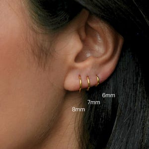 May include: Close-up of three gold hoop earrings, sized 6mm, 7mm, and 8mm, displayed on an ear. The earrings are small, circular, and shiny, reflecting light. The ear is set against a dark background.