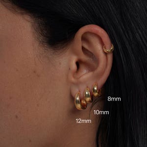 Puede incluir: Primer plano de una oreja adornada con pendientes de aro dorados. Se ven tres tamaños graduados de aros, que miden 8 mm, 10 mm y 12 mm. También se lleva un pendiente de aro dorado más pequeño en la parte superior de la oreja. Los pendientes tienen un acabado pulido.