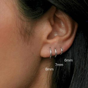 May include: Three silver hoop earrings, sized 6mm, 7mm, and 8mm, are shown on an ear. The earrings are simple, circular, and shiny. The image is a close-up shot of the earrings.