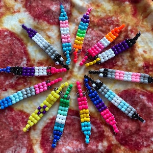May include: A collection of colorful beaded accessories, each featuring a unique pattern and color scheme. The beads are arranged in a cylindrical shape, with a loop at one end. The color combinations include rainbow, pink, blue, yellow, and black. These accessories are likely keychains or zipper pulls.
