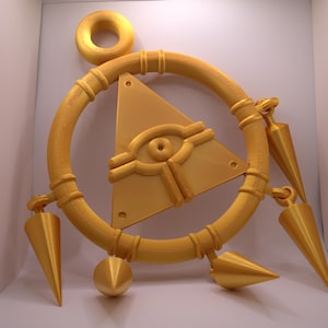 Könnte beinhalten: Goldfarbenes, 3D-gedrucktes Dekorationsobjekt mit einem kreisförmigen Rahmen und einem dreieckigen Mittelstück. Das Dreieck hat ein Augendesign. Kegel- und kugelförmige Elemente sind am Rahmen befestigt. Oben befindet sich ein Ring.