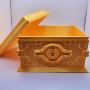 Puede incluir: Una caja amarilla con la tapa entreabierta. La caja tiene un diseño en relieve en la parte delantera, con un motivo central en forma de ojo. La caja está hecha de un material sólido y se encuentra sobre una superficie blanca.