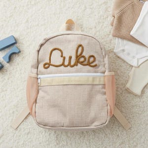 Könnte beinhalten: Ein beige-weiß gestreifter Rucksack mit dem Namen "Luke" in brauner Schreibschrift gestickt. Der Rucksack hat einen Reißverschluss, pfirsichfarbene Netztaschen und verstellbare Träger. Für Kinder konzipiert.