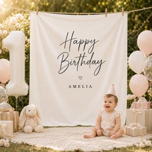 Può includere: Una bambina è seduta davanti a uno sfondo bianco con le parole "Happy Birthday Amelia" in corsivo nero. La scena comprende un palloncino con il numero uno, palloncini rosa, regali e un coniglietto di peluche. La bambina indossa un vestito rosa e un cappello da festa.