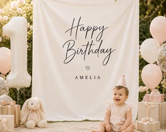 May include: A baby girl sits in front of a white backdrop with the words "Happy Birthday Amelia" in black script. The scene includes a number one balloon, pink balloons, gifts, and a stuffed bunny. The baby wears a pink dress and party hat.