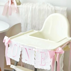 May include: A white high chair decorated with a pink "ONE" banner. The banner has scalloped edges and pink embroidered letters. Pink ribbons are tied to the high chair. A candle and pink ribbons are in the background.
