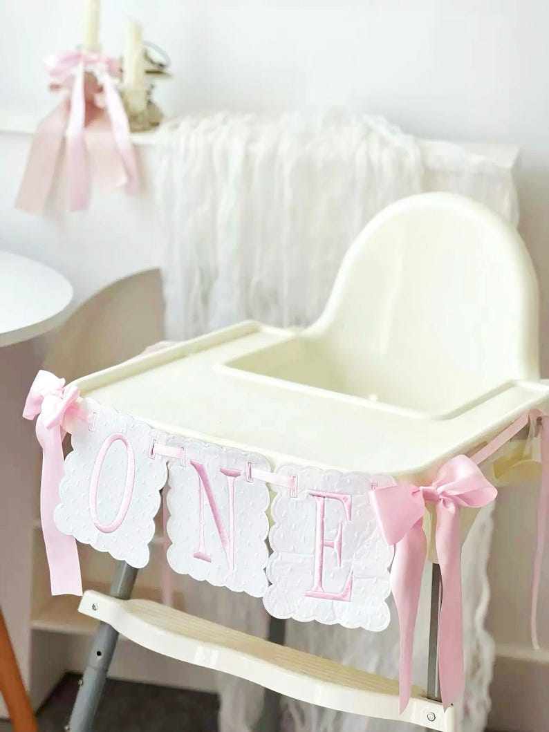 May include: A white high chair decorated with a pink "ONE" banner. The banner has scalloped edges and pink embroidered letters. Pink ribbons are tied to the high chair. A candle and pink ribbons are in the background.