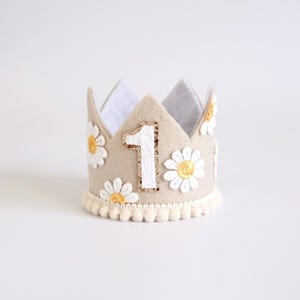May include: A beige felt crown with white daisy accents and a white number 1. The crown has a cream-colored pom-pom trim along the base. The daisies have yellow centers. A festive accessory for a first birthday celebration.