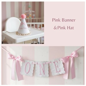 May include: A pink and white banner spelling "ONE" with pink ribbon accents, and a matching party hat with a pink pom-pom and the words "Happy Birthday". The banner and hat are set against a pink and white striped background.