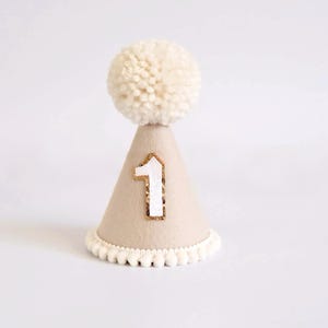 May include: A beige party hat with a white pom-pom on top and a white number 1 with gold trim. The base of the hat has a white pom-pom trim. Perfect for a first birthday celebration.