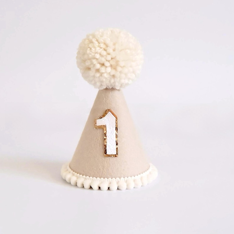 May include: A beige party hat with a white pom-pom on top and a white number 1 with gold trim. The base of the hat has a white pom-pom trim. Perfect for a first birthday celebration.