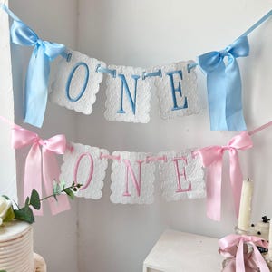 May include: Two decorative banners with the word "ONE" in blue and pink lettering. The banners are white with a scalloped edge and are adorned with matching blue and pink ribbons. The banners are likely for a first birthday celebration.