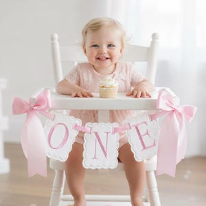 May include: A baby in a pink dress sits in a white high chair, smiling at the camera. A banner with the word "ONE" in pink letters hangs from the chair. A cupcake with white frosting sits on the tray. Pink bows adorn the sides of the banner.
